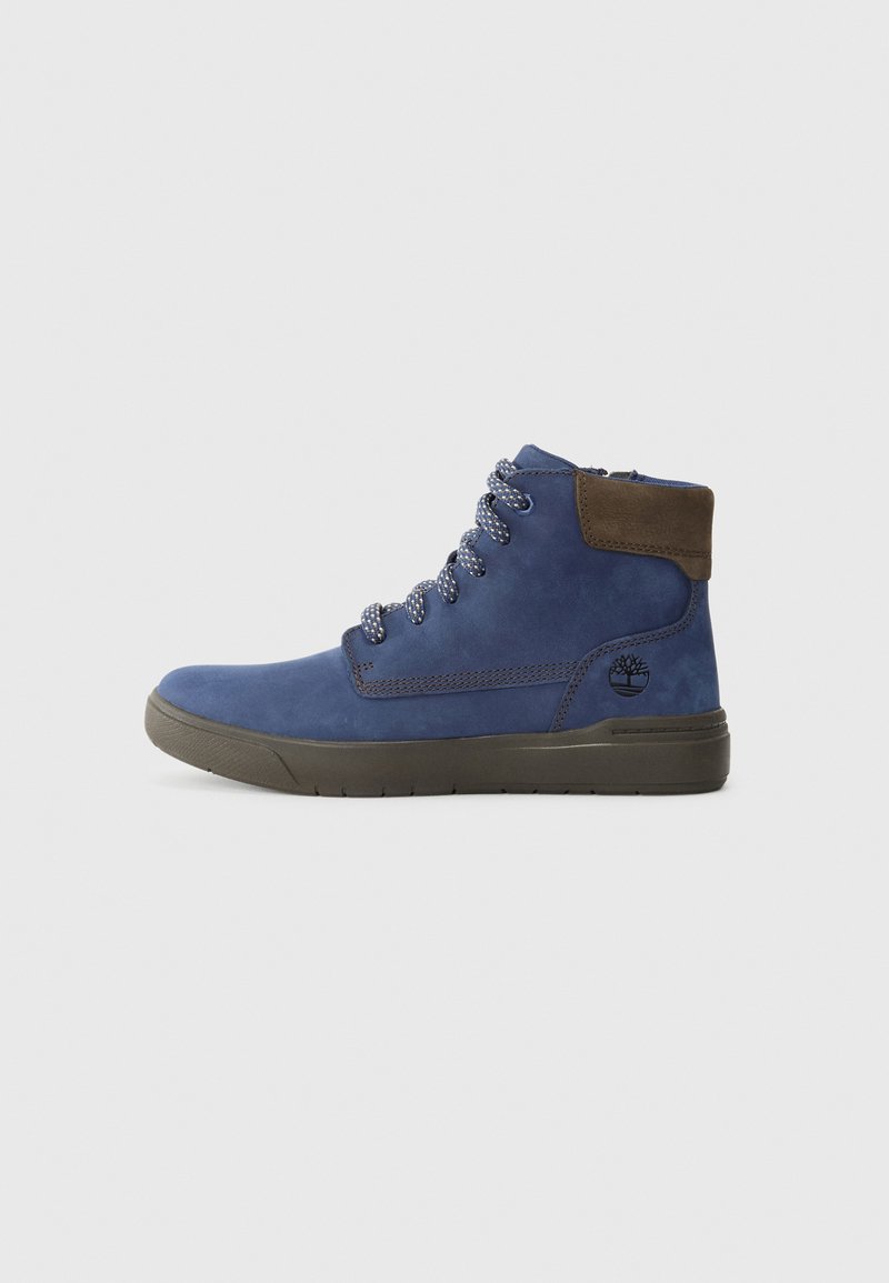 Botte montante en suede bleu avec col brun foncé, lacets à motifs, semelle noire et logo de la marque en relief sur le côté.