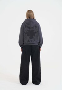 Felpa grigia oversize con una grande grafica nera sul retro, abbinata a jeans larghi neri con dettagli di ricamo colorato.