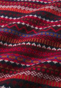 Vilnos megztinio audinys, kurį puošia raudonas, juodas ir rudanas megztas raštas su geometrinėmis formomis ir kontrastingais spalvotais dryžiais.