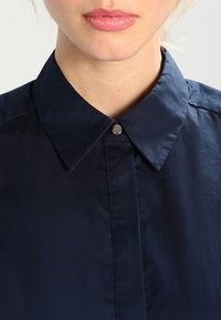 Mörkblå skjorta med krage i slät väv, med en metallisk tryckknapp vid kragen och prydlig sömnad längs den främre placketen.