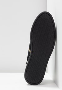 Semelle de chaussure noire avec une semelle à motif circulaire appuyée contre un mur d'angle blanc et une surface de sol.