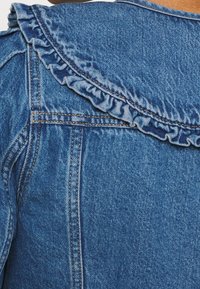 Denim jacket featuring a ruffled collar, in classic blue, with visible stitching and texture patterns typical of denim fabric.