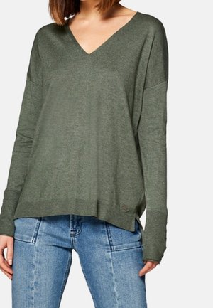 Femme portant un pull vert olive à col en V et manches longues ainsi qu’un jean bleu clair, montrée des épaules aux hanches sur fond blanc.