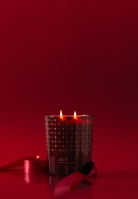 Dufte lys i en gennemsigtig sort glasbeholder med gyldne geometriske mønstre og to tændte væger, ledsaget af et rødt bånd.