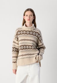 Pull en laine beige avec un col haussé à côtes, orné de motifs géométriques marron et orange complexes, au style décontracté, associé à un pantalon blanc.