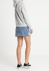 Osoba ubrana w szary hoodie, zniszczoną niebieską spódnicę dżinsową i czarne trampki, stojąca z boku na jednolitym białym tle.