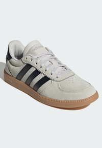 Siva sportska cipela od brušene kože s tri crne pruge, svijetlosivim vezicama i potplatom boje gume. Ima podstavljeni okovratnik i prozračne perforacije na vrhu za ventilaciju.