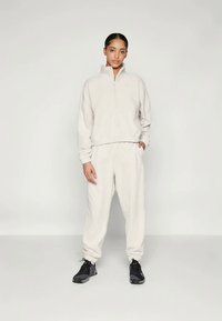 Beige fleecegenser og matchende bukser, glidelås-krage, sidelommer, elastiske mansjetter, kombinert med svarte treningssko. Glatt tekstur.