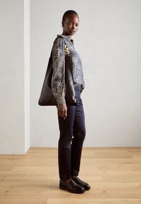 Svart skinn tote-veske med en glatt tekstur. Modellen har på seg en marineblå trykt bluse, marineblå skinny bukser og svarte loafers, stående på tregulv.