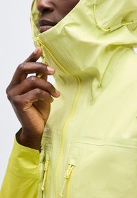 Άτομο που φορά ένα ανοιχτό κίτρινο μπουφάν με κουκούλα, τραβώντας το φερμουάρ μπροστά κοντά στο στόμα, δείχνοντας τις λεπτομέρειες του υφάσματος και των κλειστών του μπουφάν.