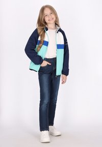 Ombygbar jakke i marineblå, mint og hvid med lynlås; parret med blå jeans og hvide sneakers. Lige silhuet, blød tekstur.