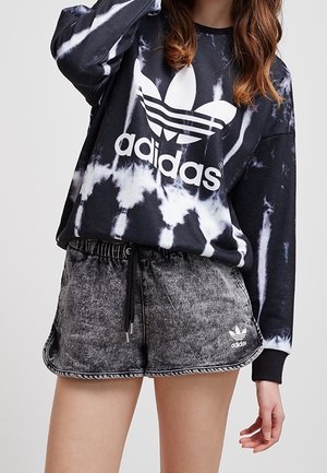Osoba ubrana w czarno-białą bluzę Adidas tie-dye i szare spodenki Adidas ze sznurkiem w pasie, stojąca na jednolitym tle.