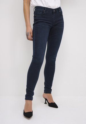 Kvinde iført mørkeblå skinny jeans og sorte spidse højhælede sko, stående mod en ensfarvet lys baggrund.