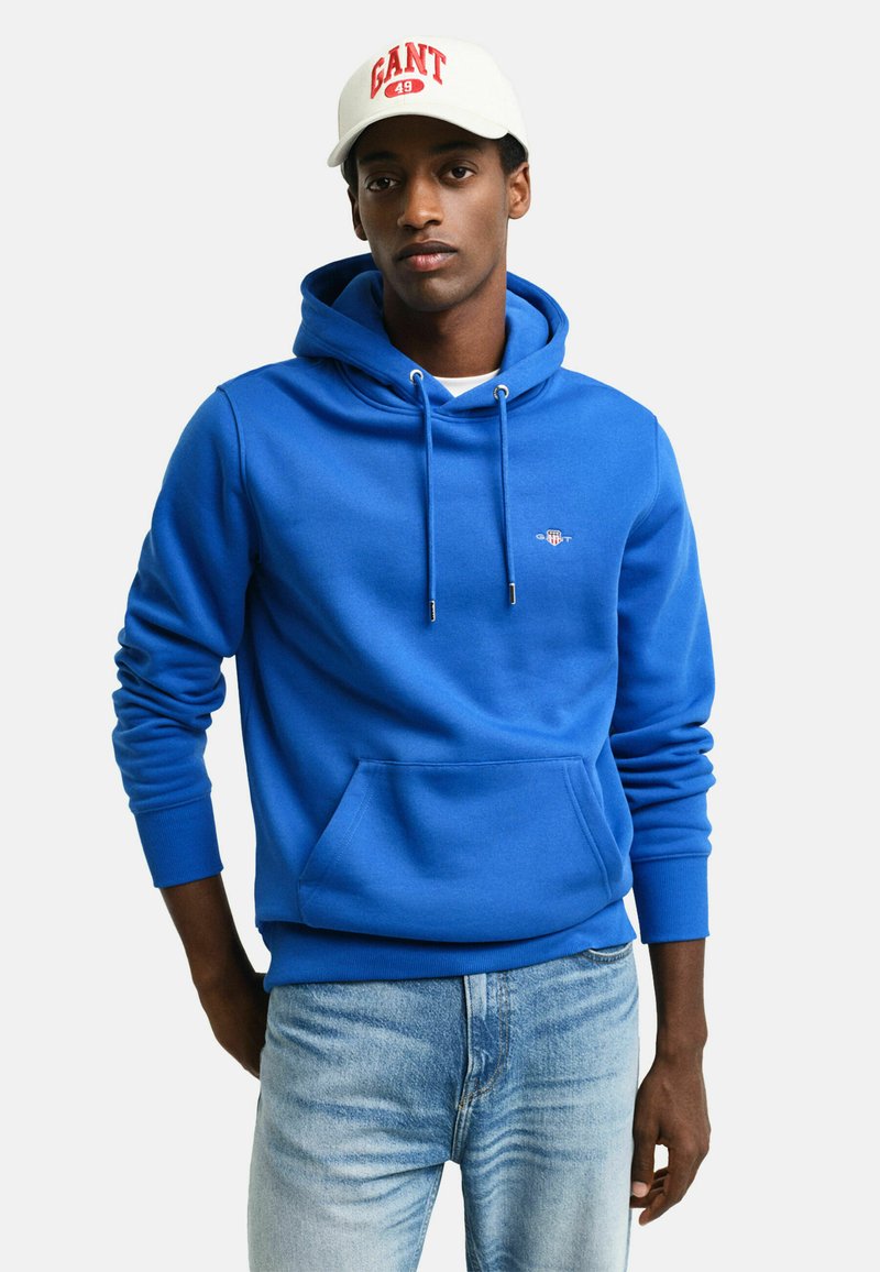 Blauwe hoodie met een voorzak, gemaakt van zacht materiaal. Bevat verstelbare trekkoorden en een klein logo op de borst.