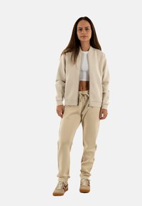 Chaqueta bomber beige sobre un top corto blanco, combinada con pantalones de chándal beige y zapatillas marrones. Tela suave, corte relajado y cintura ajustable.