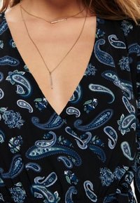 Tmavé oblečení s paisley vzorem v modré a teal barvě se květinovými akcenty. Hluboký výstřih doplněný dvěma vrstvenými náhrdelníky.