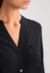 Blouse noire à col en tissu lisse. Comprend une poche plaquée, une fermeture boutonnée à l'avant et une coupe décontractée. Design simple avec des accents subtils.