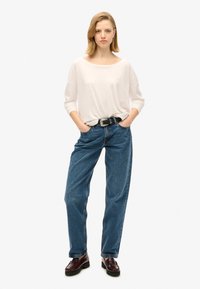 Lichtbeige langemouwen top, losse pasvorm; blauwe hoog-waisted jeans; zwarte riem met zilveren gesp; donkerbruine loafers.