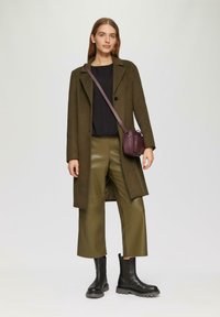 Manteau vert olive avec un col cranté, haut noir texturé, pantalon en cuir olive raccourci et bottes à semelle épaisse noires. Sac bandoulière bordeaux.
