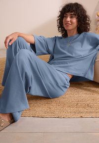 Ensemble de vêtements d'intérieur bleu clair en matière douce, comprenant un haut ample à manches courtes et un pantalon à taille élastique. Tapis texturé au sol en dessous.