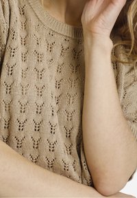 Primo piano di una persona che indossa un maglione lavorato a maglia beige con un motivo a foglia testurizzato. Una mano è appoggiata vicino al viso, con le dita che toccano delicatamente.