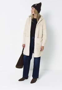 Cappotto in finta pelliccia crema con due tasche frontali, cappuccio e zip, abbinato a jeans di denim scuro e un berretto lavorato a maglia marrone. Borsa marrone e stivaletti.