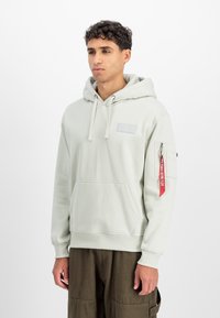 Ανοιχτό γκρι φούτερ με τσέπη καγκουρό, κορδόνια και ένα πατσιόν. Διαθέτει τσέπη με φερμουάρ στο μανίκι με κόκκινη ετικέτα και ριχτό τελείωμα.