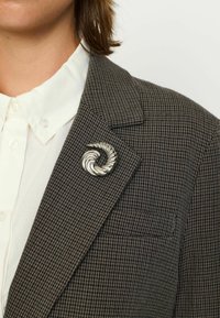 Spilla a spirale argento su un blazer in tessuto scozzese marrone con una camicia bianca con colletto sotto, caratterizzato da rever affilati e una tasca.