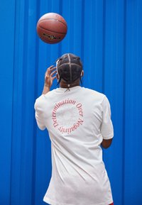 Valkoinen t-paita, jossa on punaiset kaarevat tekstit "Pyrkimys Yli Negatiivisuuden" selässä, yhdistettynä koripallon kanssa sinistä taustaa vasten.