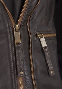 Ruskea nahkatakki, jossa on kultaiset vetoketjut, mukaan lukien etupocketin vetoketju. Takissa on teksturoitu pinta ja näkyviä ompeluyksityiskohtia.