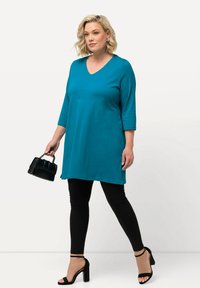 Teal V-ringad tunika med trekvartslånga ärmar, ihop med svarta leggings och remmade högklackade sandaler, hålla en liten svart väska.
