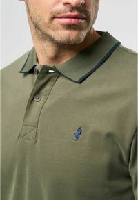 Roheline polo särk tumesinise kraega, kahe nupuga ja väikese sinise tikandi logoga. Materjal näib olevat pehme ja sile tekstuur.