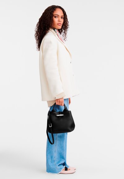 Schwarze Lederhandtasche mit strukturierter Oberfläche, silbernem Zubehör und kurzen Tragegriffen. Getragen von einem Model in einem creamfarbenen oversized Blazer und blauen Jeans.