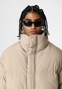 Beige pufferjack met hoge kraag, gestructureerde stof en naaideetails. Draagt een paar zwarte zonnebril.