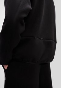 Čierny pulóver s textúrou a priliehavým strihom, ktorý má akcent na leme a diskrétnu bočnú kapsu. Švy na rukávoch pridávajú dizajnový záujem.