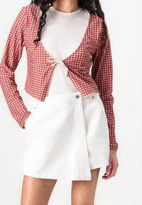 Top corto de cuadros rojo y blanco con mangas largas, atado al frente con un cordón crema; combinado con una falda mini de mezclilla blanca.