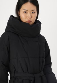 El abrigo de plumas negro presenta un alto collar plegable, cintura ceñida con cinturón y textura acolchada, ofreciendo calidez y una silueta estructurada.