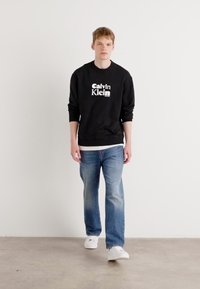 Black sweatshirt with white "Calvin Klein" logo, paired with light blue jeans and white sneakers; smooth fabric, relaxed fit, standard crew neck.