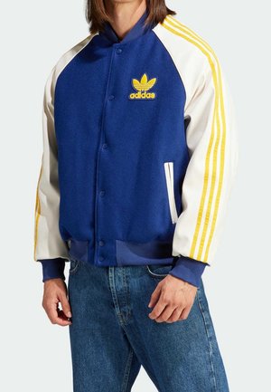 Person trägt eine blau-weiße Adidas-Varsityjacke mit gelben Streifen an den Ärmeln und blaue Jeans, steht mit entspannten Händen.