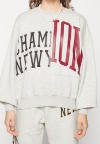 Felpa grigia oversized con grande stampa "CHAMPION" e "NEW YORK" in nero e bordeaux, dal taglio rilassato e con polsini a costine.