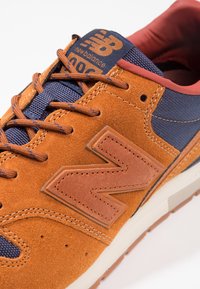 Oranžové semišové běžecké boty s námořnickými síťovanými detaily, velkým hnědým logem New Balance, hnědými tkaničkami a bílou gumovou podrážkou.