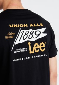 T-shirt en coton noir avec un texte graphique doré et blanc. Le texte comprend "UNION ALLS," "1889," "Vêtements de travail durables," et "Lee."