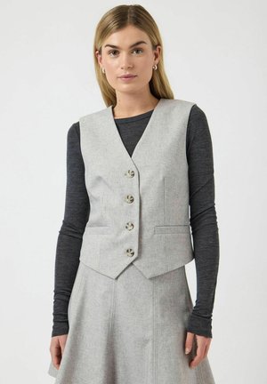 Young woman with straight blonde hair wearing a light gray buttoned vest and matching skirt over a long-sleeve dark gray top.