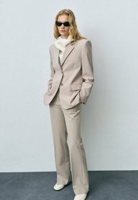 Costume beige ajusté avec un blazer structuré et un pantalon évasé, associé à un pull col roulé blanc en maille et des lunettes de soleil noires. Finition en tissu lisse.