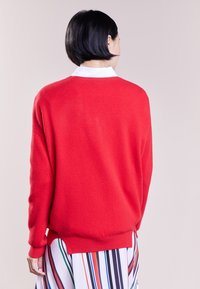 Suéter rojo hecho de un material suave con un corte relajado, que presenta un dobladillo y puños acanalados. Lleva puesto sobre una falda de colores a rayas.