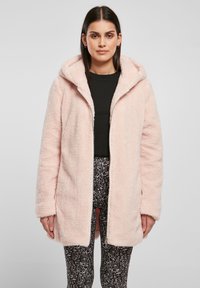 Cappotto in faux fur rosa chiaro con cappuccio, chiusura con zip e tessuto texturizzato. Indossato sopra un top nero e leggings con fantasia.