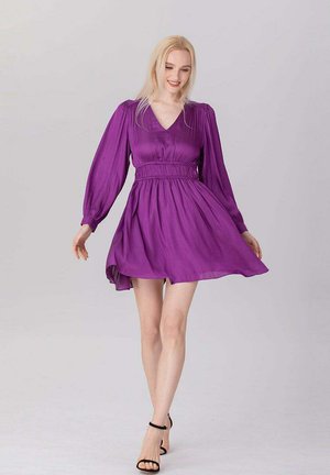 Femme en robe longue fluide violette à manches longues et escarpins noirs avec bride à la cheville, debout sur un fond clair uni, souriante et regardant vers le bas.