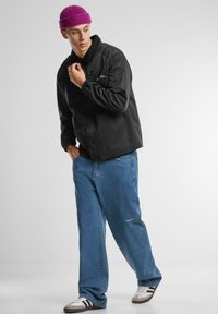 Black lightweight jacket with front buttons, paired with light blue baggy jeans, white sneakers with black stripes, and a purple knit beanie.