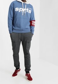 Blå huvtröja med vit "sports" text och röd ärmaccent, i kombination med grå fleece joggers och röda sneakers med vita ränder.