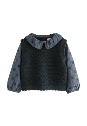 Pull sans manches tricoté vert foncé porté sur une blouse grise à manches longues avec des motifs floraux découpés et un col à volants.